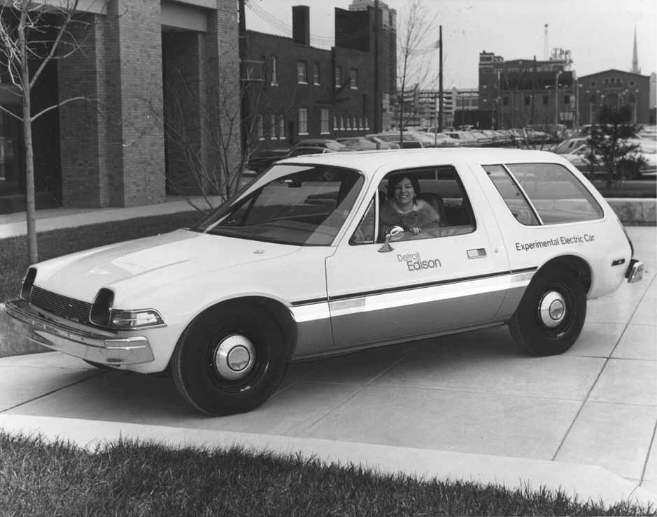 Photo noir et blanc d'une femme souriante se penchant hors du siège du conducteur d’une compacte familiale. Texte sur le côté du véhicule : Detroit Edison / Voiture électrique expérimentale.