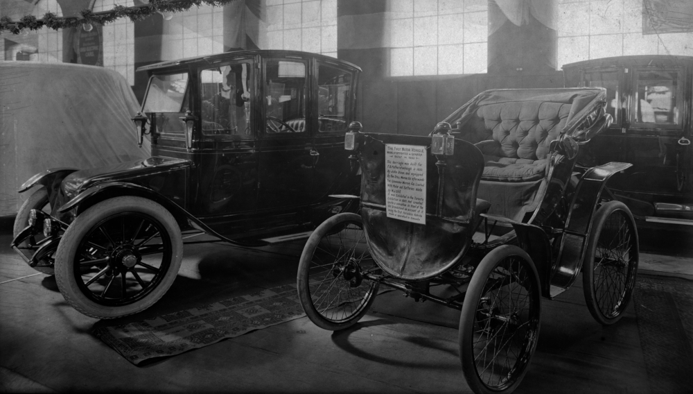 Deux voitures électriques l'une à côté de l'autre dans un espace d'exposition. Le panneau sur la plus petite indique :  PREMIER VÉHICULE À MOTEUR .