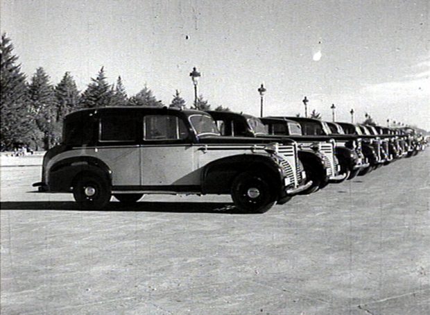 Image noir et blanc de plusieurs automobiles garées côte à côte dans un parc de stationnement vide avec, à l’arrière-plan, des pins et des lampadaires décoratifs.