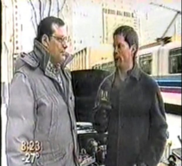 Image montrant deux hommes en gros manteaux d'hiver devant une voiture au capot ouvert dans une rue de la ville, avec un tramway passant en arrière-plan. L'homme à droite tient un microphone. Texte en bas à gauche de l’écran : 8:23 -28°