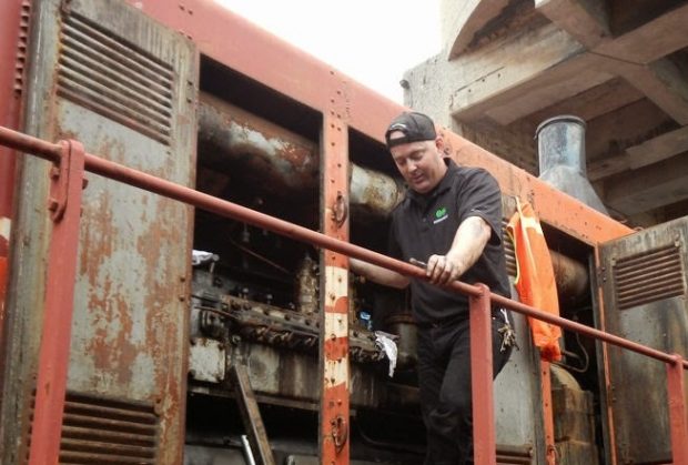 Photographie couleur d'un homme debout sur la plate-forme d'une locomotive avec les portes du moteur ouvertes. Il regarde vers le bas et a un outil à la main.