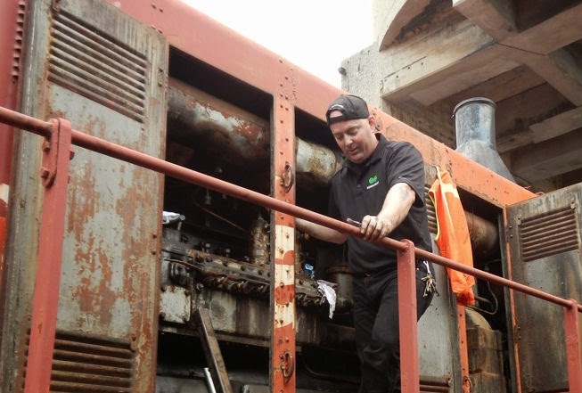 Photographie couleur d'un homme debout sur la plate-forme d'une locomotive avec les portes du moteur ouvertes. Il regarde vers le bas et a un outil à la main.