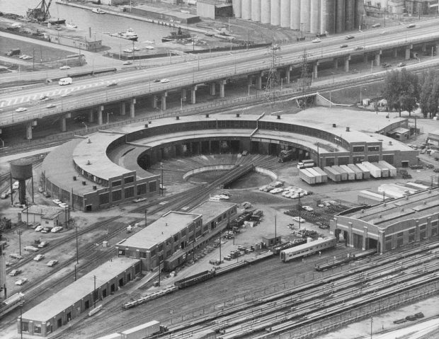 Photographie aérienne d’époque en noir et blanc montrant une rotonde et des voies ferrées. Il y a une autoroute à l’arrière-plan et deux bâtiments rectangulaires au premier plan.