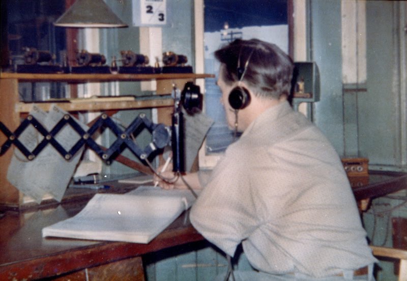 Un homme est assis sur son bureau et écrit dans un grand livre. Il porte un casque d’écoute et un téléphone d’époque est tendu vers lui.
