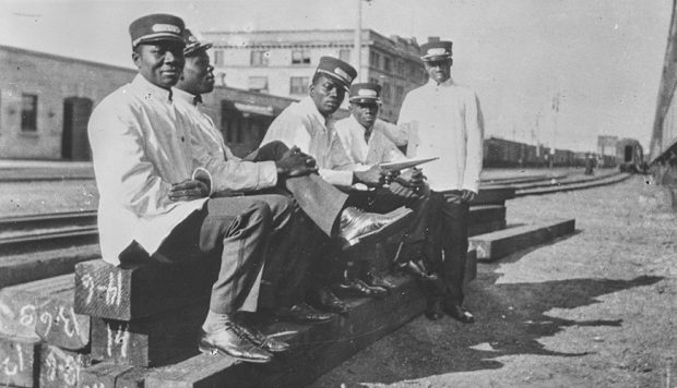Photographie d’archives en noir et blanc de cinq hommes noirs assis sur une voie ferrée. Ils sont vêtus d’uniformes ferroviaires blancs.