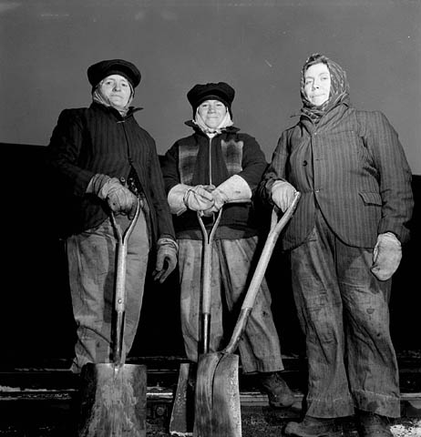 Photographie d’archives en noir et blanc de trois femmes posant avec des pelles. Elles sont vêtues de manteaux et de chapeaux, d’écharpes et de gants de travail. Elles regardent l’appareil photo.