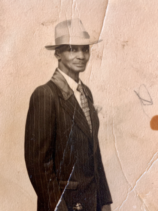 Photographie d’archives en noir et blanc aux tons sépia d’un homme partant de la taille. Il porte un chapeau et un costume et sourit légèrement.
