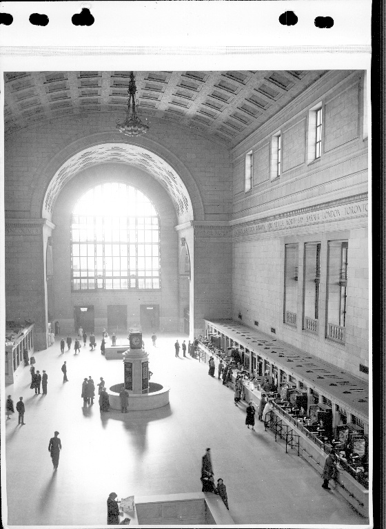 Photographie d’archives en noir et blanc de l’intérieur d’une grande gare ferroviaire. La photographie est prise d’en haut et montre de nombreux guichets sur le côté droit. Il y a plusieurs personnes qui marchent dans la gare.