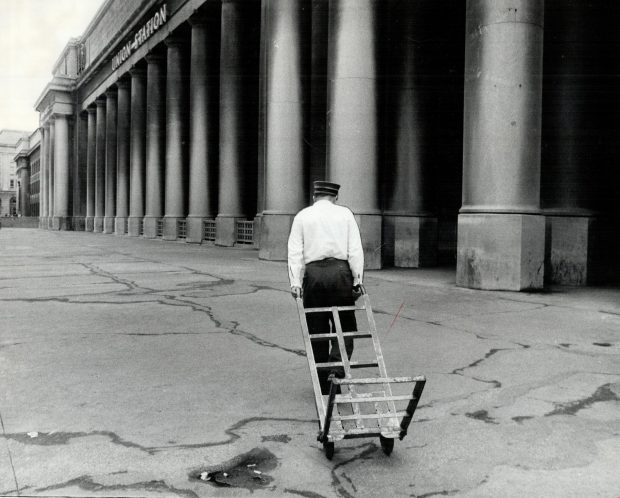 Photographie d’archives en noir et blanc d’un homme en uniforme de cheminot tirant un chariot à bagages vide devant la gare Union de Toronto. Il s’éloigne de l’appareil photo.
