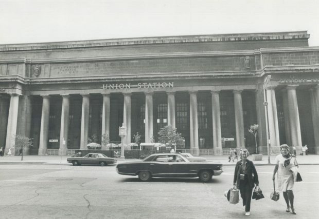 Photographie d’archive en noir et blanc de la façade d’une grande gare ferroviaire. On aperçoit des voitures d’époque circulant dans la rue devant la gare et deux femmes portant des sacs à provisions au premier plan.