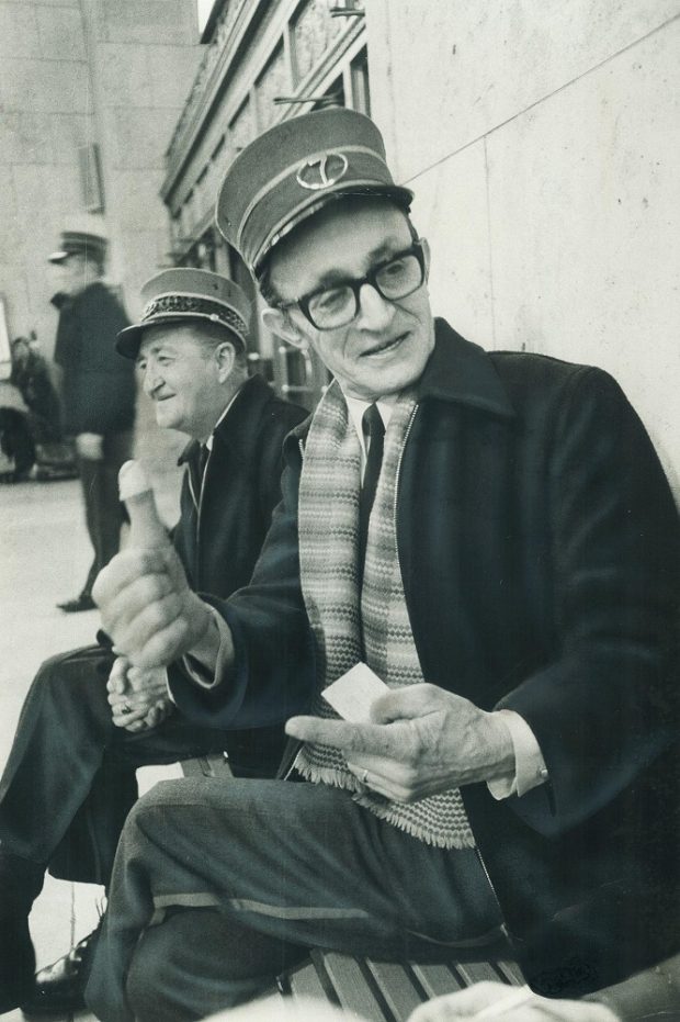 Photographie d’archives en noir et blanc de deux hommes assis sur un banc. Les hommes portent des uniformes ferroviaires, y compris des chapeaux. L’homme au premier plan parle et fait des gestes avec ses mains. L’homme à l’arrière-plan détourne le regard de l’appareil photo.