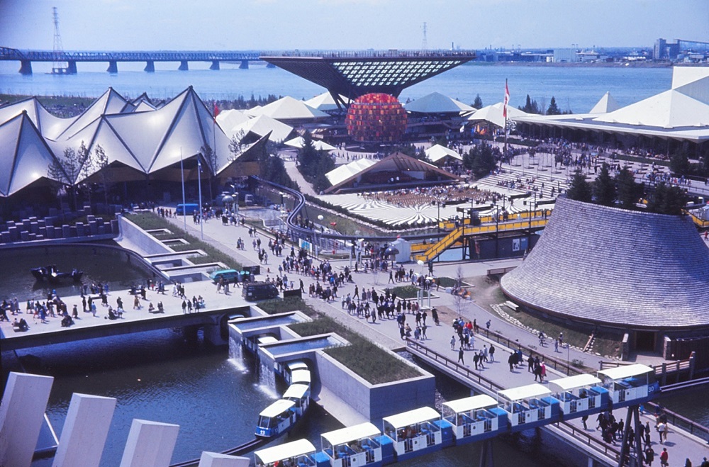 Photographie d’archive en couleur d’Expo 67. La photographie est prise d’en haut et montre quatre bâtiments courts aux formes inhabituelles. De nombreuses personnes marchent sur les routes entre les bâtiments.
