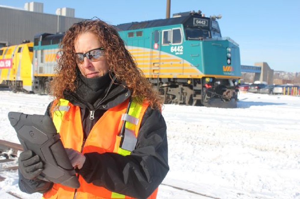 Photographie moderne en couleur d’une femme portant un gilet de haute visibilité travaillant sur une tablette. On aperçoit une grande locomotive verte à l’arrière-plan.