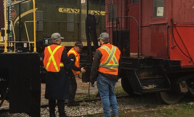 Photographie couleur de trois hommes portant des gilets de haute visibilité se tenant entre une locomotive et un fourgon de queue. Deux des hommes sont tournés vers l’extérieur de l’appareil photo. L’homme au centre est tourné vers la caméra et pousse un coupleur sur le fourgon de queue.