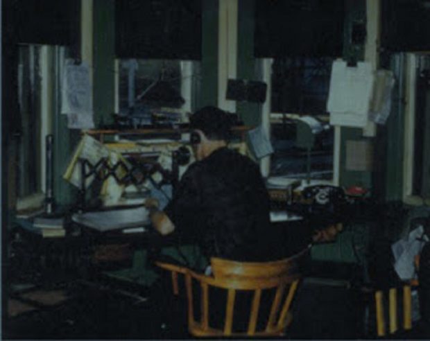 Une photographie d’époque d’un homme travaillant à un bureau. Il est vu de dos. Il porte un casque et un téléphone d’époque est tiré vers lui. Il y a beaucoup de papiers sur le bureau.