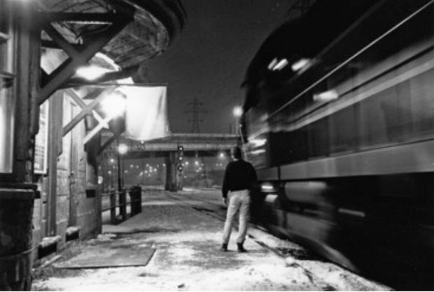 Un homme se tient sur un quai de gare tandis qu’un train passe sur la droite. Il tient un grand bâton en direction du train. On peut voir une partie de la gare sur le côté gauche.