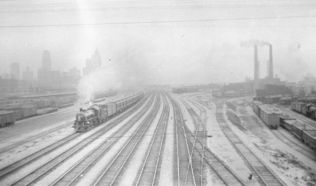 Photographie d’époque en noir et blanc montrant des voies ferrées. Une locomotive à vapeur se trouve sur le côté gauche et la vapeur s’élève vers le haut. L’arrière-plan est obscurci par le brouillard.