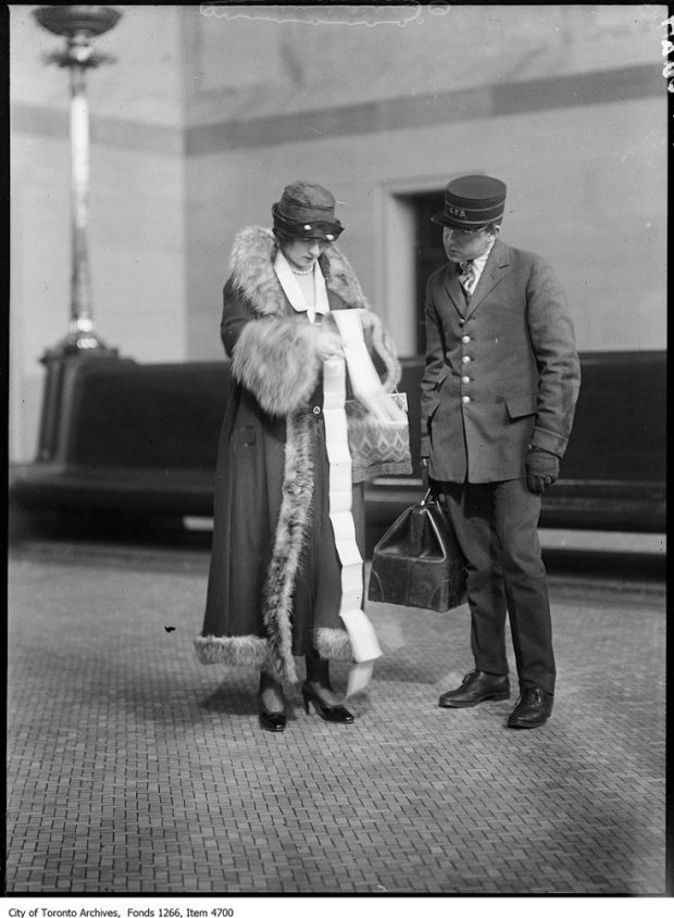 Photographie d’archives en noir et blanc d’une femme richement vêtue et parcourant un long billet de train tandis qu’un homme en uniforme regarde également le billet.