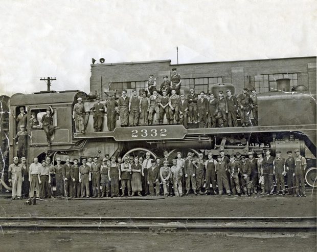 Environ 75 personnes posent devant et sur une grande locomotive à vapeur posée sur des voies ferrées. Le numéro de locomotive « 2332 » est visible au centre de la locomotive. Une autre série de voies ferrées vides se trouve au premier plan et un court bâtiment rectangulaire à l’arrière-plan.