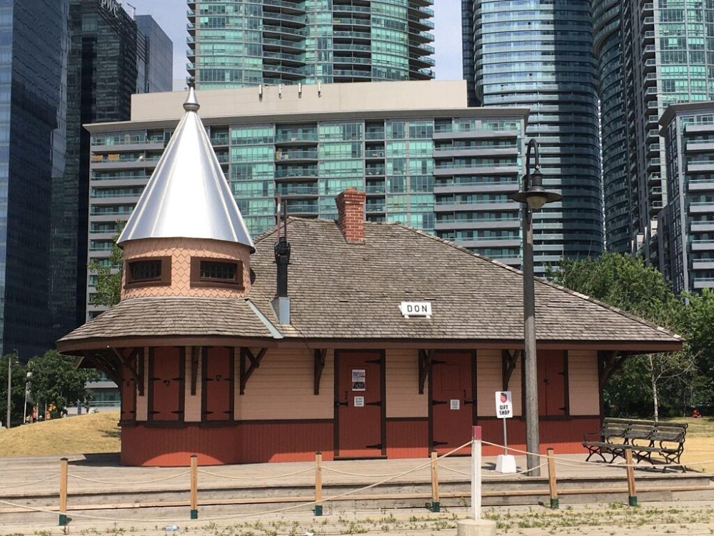 Une photographie moderne d’une petite gare avec des cordes et des rails de train miniatures au premier plan. « DON » est écrit sur le toit de la gare et il y a un petit panneau « BOUTIQUE DE SOUVENIRS » devant la gare.