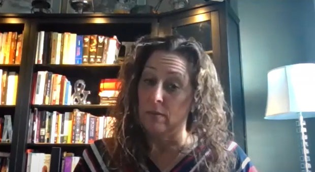 Capture d'écran couleur d'une femme regardant vers le bas. La tête et les épaules sont visibles, avec des étagères de livres en arrière-plan.