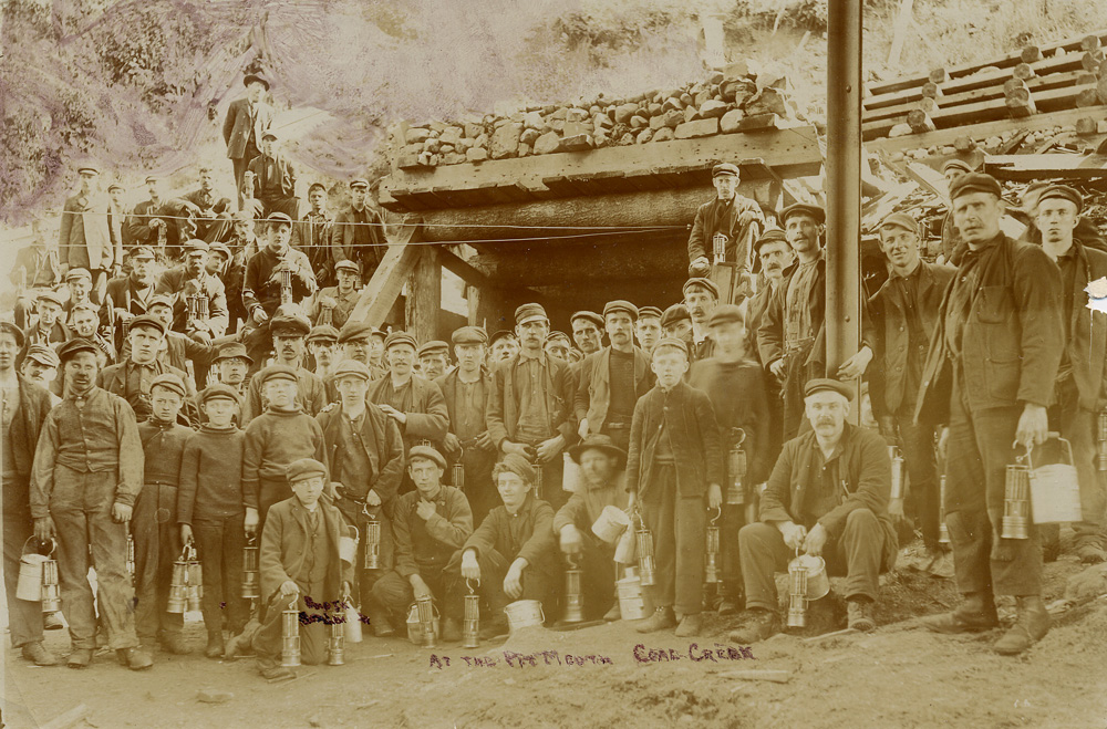 Groupe de mineurs tenant des lampes de mine et des seaux à lunch devant une entrée de mine.