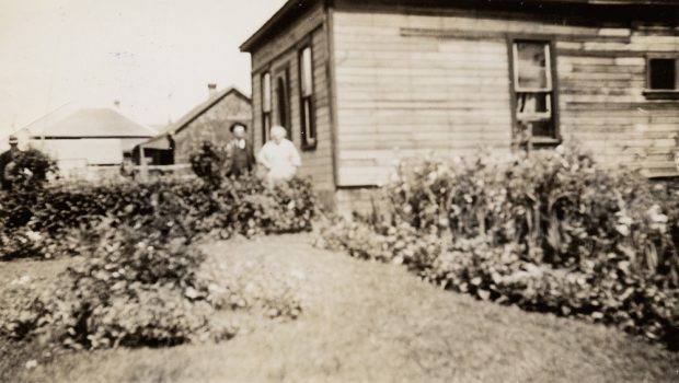  Un homme âgé et une femme debout près de la porte d'entrée de leur maison; un grand jardin entoure la maison.