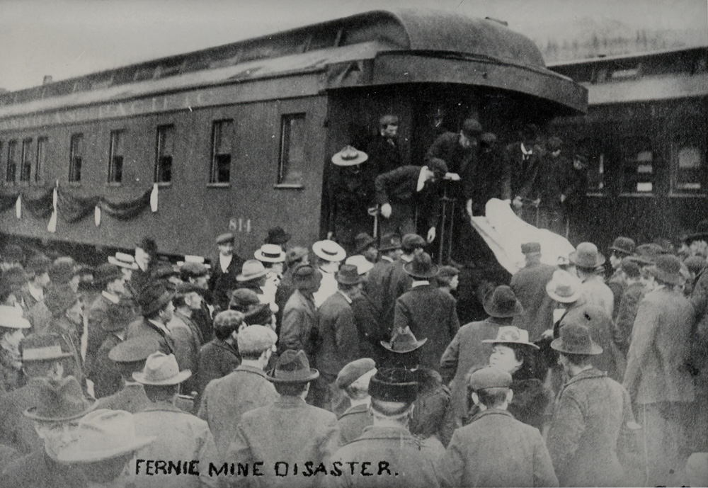 Grande foule d'hommes et de femmes devant une voiture de chemin de fer. Un cadavre recouvert d'une bâche est déchargé d'une voiture de train.