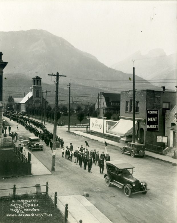  Une voiture mène une grande procession funéraire. Les hommes marchent à deux derrière la voiture.