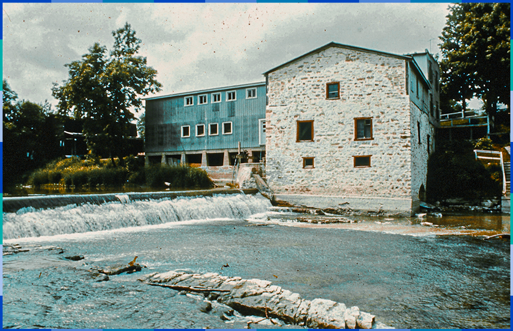 Photographie en couleurs présentant la face sud du moulin Légaré. On y voit la digue, le moulin en pierres et la scierie qui y est connectée perpendiculairement.