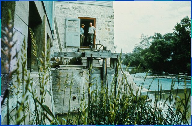La photo présente la face ouest du moulin et est prise au niveau de l’eau. Donat et Philippe Légaré sont debout, au haut des marches qui mènent à la porte d’eau.