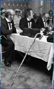 Photographie noir et blanc de trois conseillers assis au coin d’une table qui rigolent. Des chapeaux haut-de-forme, des cannes et une lampe à l’huile sont déposées sur la table.