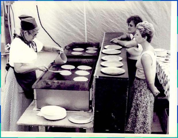 Photographie en noir et blanc de deux femmes qui se tiennent derrière un comptoir et regardent une cuisinière qui verse le mélange à galette sur une plaque chauffante. Plusieurs assiettes garnies de galette sont déposées sur le comptoir.