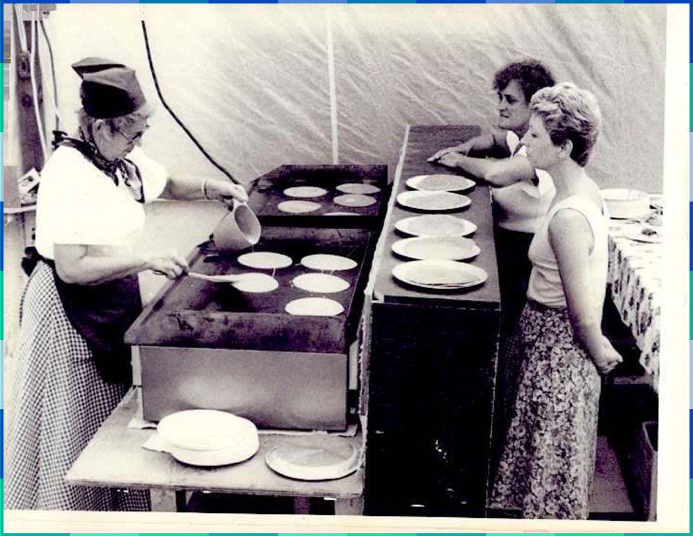 Photographie en noir et blanc de deux femmes qui se tiennent derrière un comptoir et regardent une cuisinière qui verse le mélange à galette sur une plaque chauffante. Plusieurs assiettes garnies de galette sont déposées sur le comptoir.