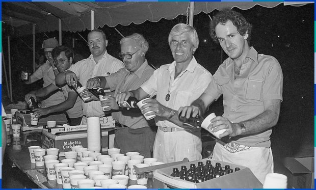 Photographie noir et blanc de six hommes qui posent s’affairent à décapsuler des bouteilles de bière pour verser le liquide dans de petits verres en carton. Devant eux, une table où sont déposés une centaine de verres et plusieurs boîtes de bières.
