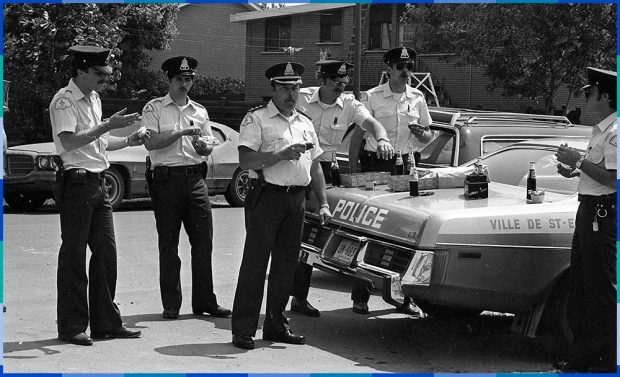 Photographie noire et blanc de six policiers réunis autour d’une voiture de patrouille. Ils mangent des beignes et boivent du Pepsi en bouteille de vitre.