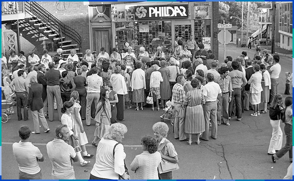 Une vingtaine de femmes, certaines jeunes et d’autres âgées, sont assises en file le long de la rue Saint-Eustache pour tricoter. Devant elles, une foule d’une cinquantaine de personnes les regarde.