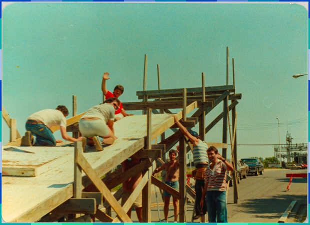 Six hommes du Club Optimiste saluent la caméra durant la construction de la rampe en bois. Celle-ci mesure environ trois mètres de haut. 