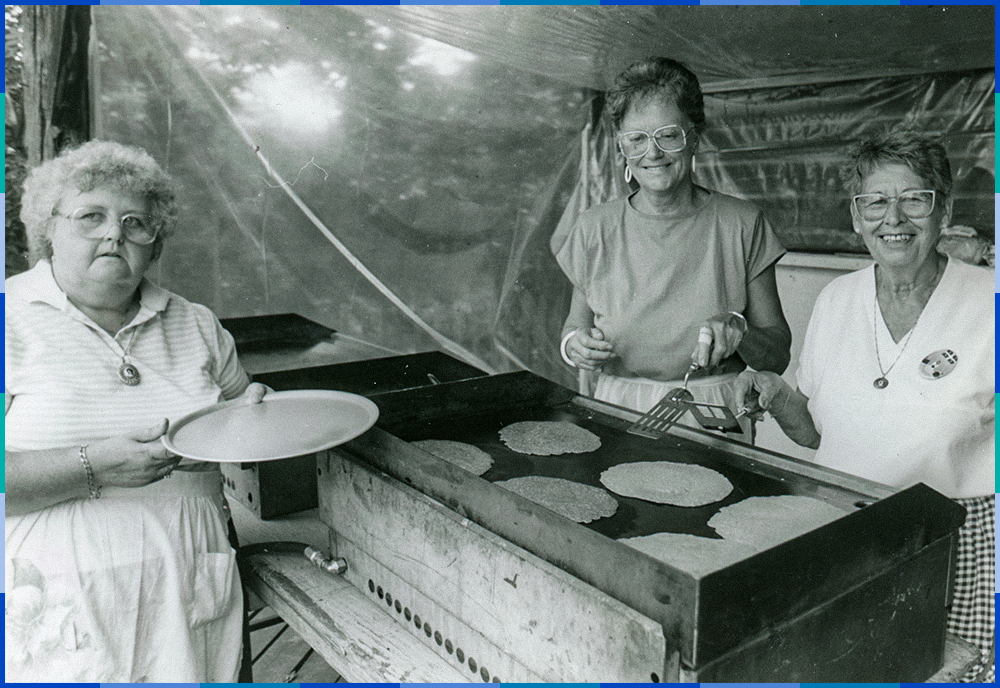 Deux cuisinières préparent des galettes sur une plaque de cuisson tandis qu’une femme attend, assiette à la main.