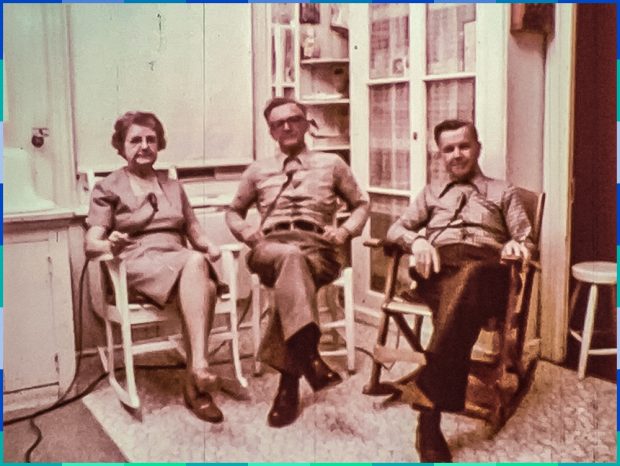 Une femme et deux hommes âgés sont assis sur des chaises berçantes ou droite dans une cuisine. Ils regardent la caméra. 