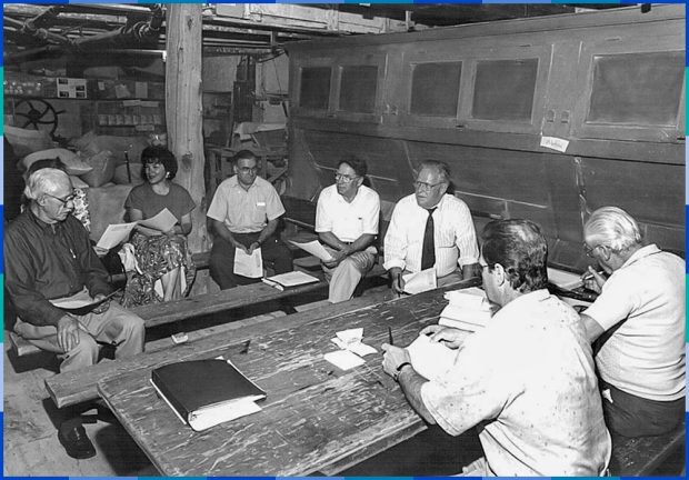 Les huit membres du conseil d’administration sont assis autour d’une table de bois à l’intérieur du moulin Légaré. À droite de la photo, on observe le bluteau qui sert à séparer la farine du son.