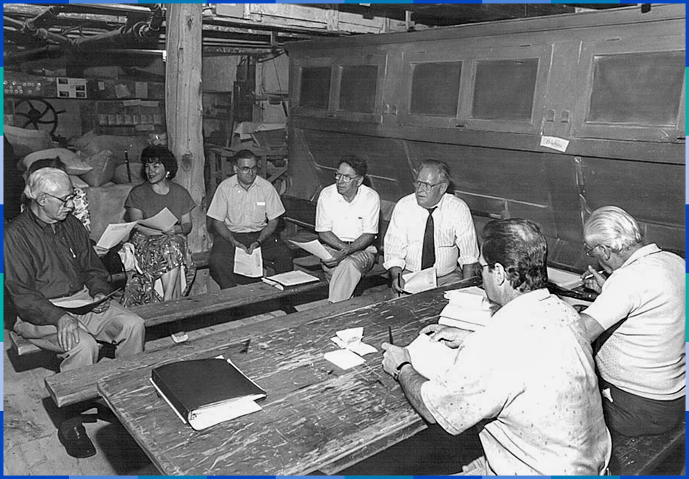 Les huit membres du conseil d’administration sont assis autour d’une table de bois à l’intérieur du moulin Légaré. À droite de la photo, on observe le bluteau qui sert à séparer la farine du son.