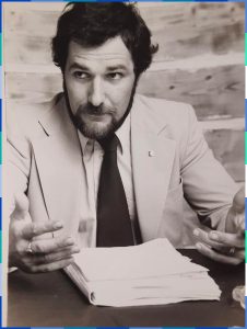 Photographie en noir et blanc d’un homme barbu en habit avec cravate. Il est assis à un bureau et une pile de documents est posée devant lui.