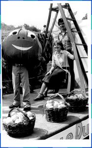 Photographie en noir et blanc d’un homme debout et une femme assise sous un escabeau se faisant prendre en photo avec une personne déguisée en pomme souriante. Devant eux sont placés trois paniers remplis de pommes.