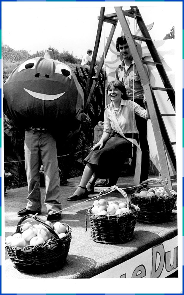 Photographie en noir et blanc d’un homme debout et une femme assise sous un escabeau se faisant prendre en photo avec une personne déguisée en pomme souriante. Devant eux sont placés trois paniers remplis de pommes.