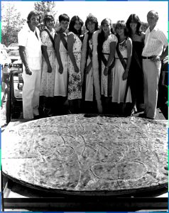 Photographie en noir et blanc de sept jeunes femmes et deux hommes qui prennent la pose derrière une énorme tarte aux pommes sur laquelle l’inscription ‘’Festival de la pomme’’ est gravée. 