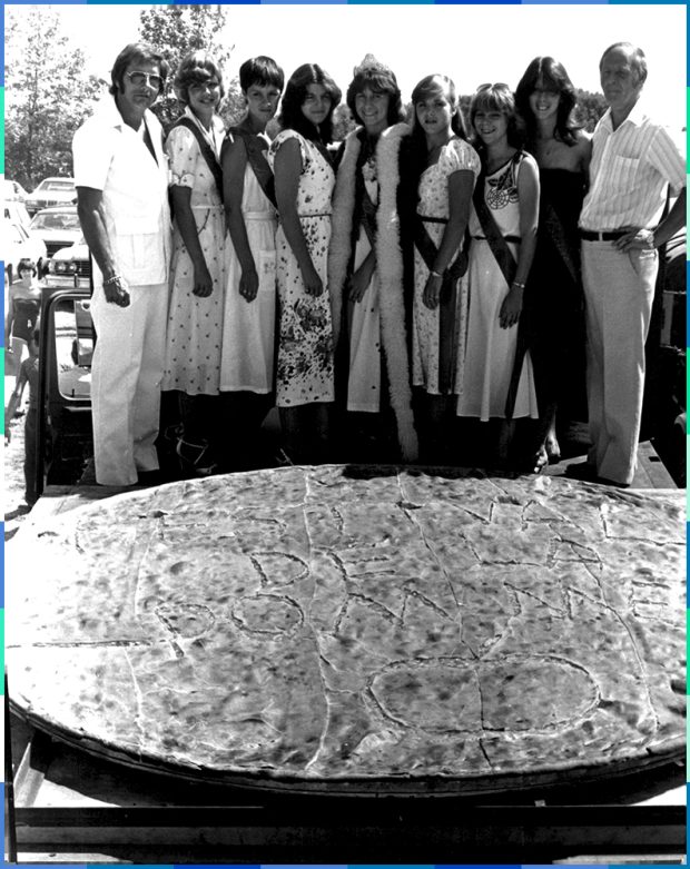 Photographie en noir et blanc de sept jeunes femmes et deux hommes qui prennent la pose derrière une énorme tarte aux pommes sur laquelle l’inscription ‘’Festival de la pomme’’ est gravée. 