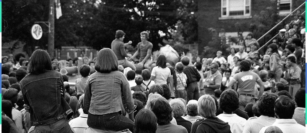 Photographie en noir et blanc d’une foule réunie dans la rue autour de deux hommes assis et surélevés. Deux jeunes filles sont assises sur les épaules d’une autre personne.  