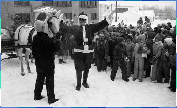 Photographie en noir et blanc d’un homme déguisé en père Noël qui rend visite à des enfants dans la cour d’une école primaire. Ses cadeaux sont placés dans une calèche tirée par un cheval blanc. 