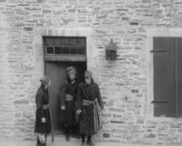 Trois patriotes portant des tuques et des longs manteaux fermés grâce à des ceintures fléchés sont devant la porte d'un bâtiment de pierres.